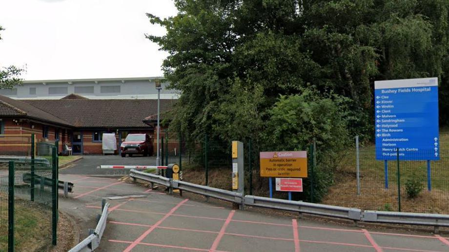 Entrance to Bushey Fields Hospital showing blue NHS signs, trees and grass areas, red chevrons on the driveway and buildings in the background.