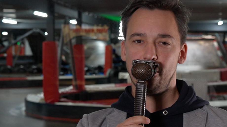 Alex Jacques, holding a lip mic, in front of a karting track. He is wearing a grey blazer, over a black hoodie.