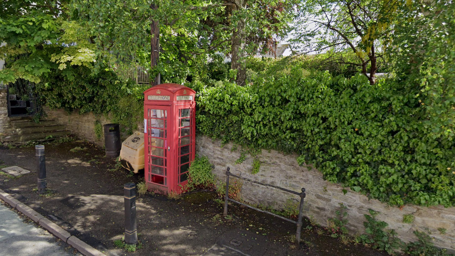 A red phone box on a pavement. There is a brick wall behind it with green bushes and trees