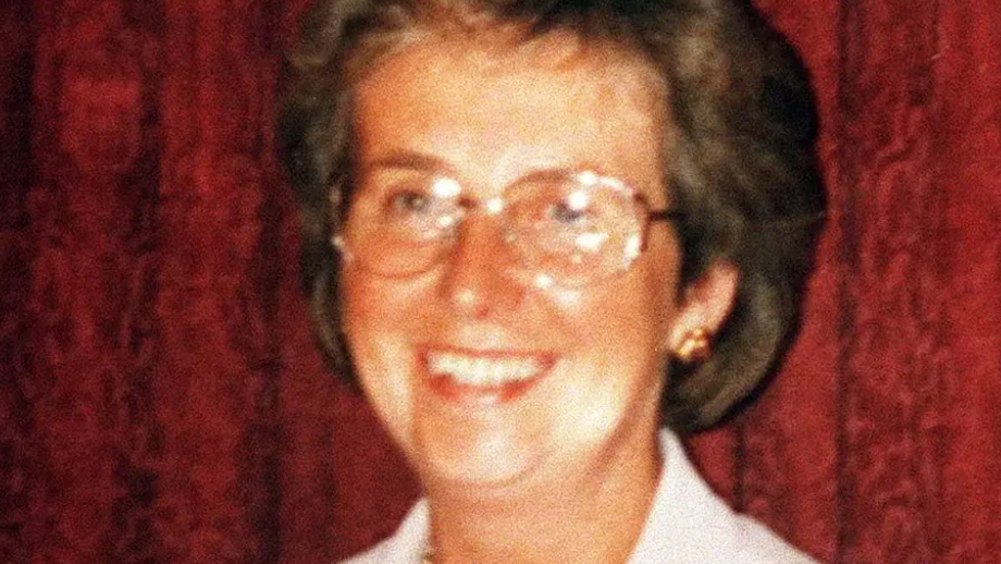 Woman with short brown hair and large round glassed standing in front red curtain