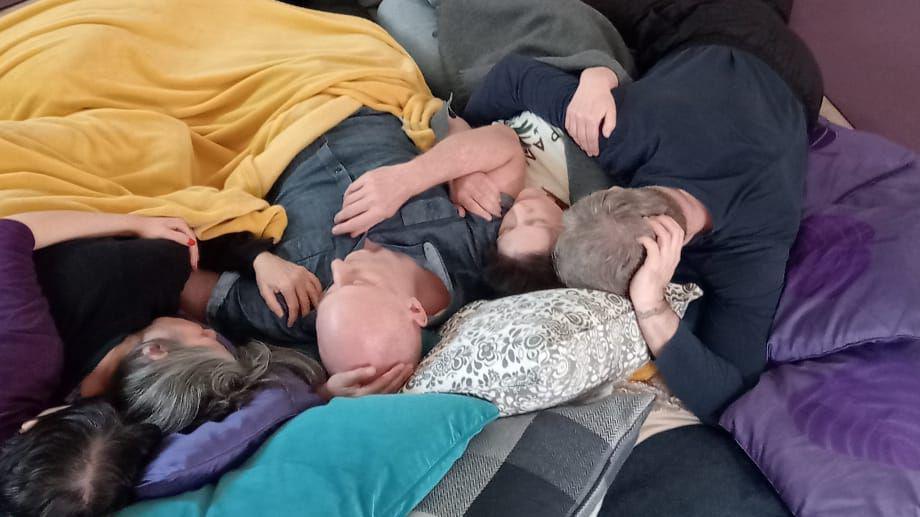 A group of people are covered in blankets and united in cuddles as they lie on a nest of cushions. No faces are clearly visible but there are men and women of various ages.