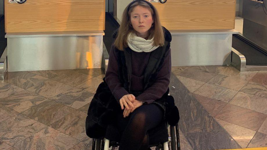 A woman with brown hair, dressed in black with a white scarf, sits in a wheelchair in front of an airport check-in desk 