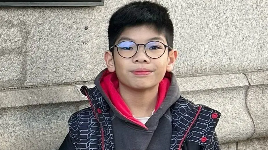 Thomas Wong stares directly at the camera. He has large glasses on his face and dark hair. He is wearing a grey hoodie with red interior lining and a black jacket with red zip and button and a white box design. He is standing in front of a light stone wall.