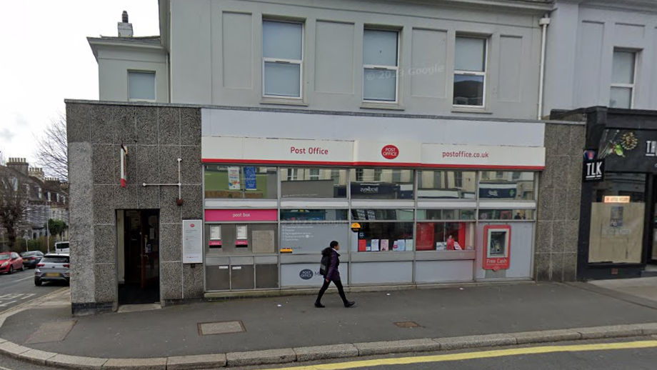 Photo of the Mutley Post Office, a grey building on a street corner, with a woman walking in front on the pavement