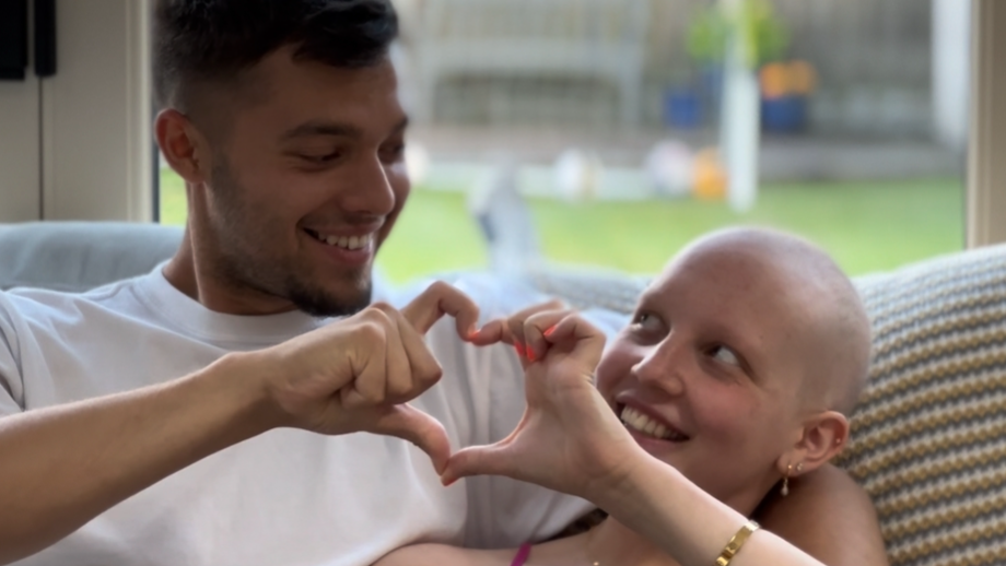 Izzy Fletcher and her boyfriend make a heart symbol with their joined hands as they sit smiling at each other on a sofa. She is bald while her boyfriend has dark hair shaved at the sides and a short beard which is longer on the chin.