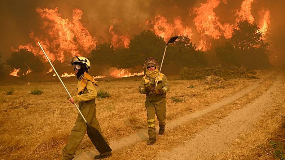 Two female firefighters tackle a raging inferno in a Spanish woodland scene. The grass around them is dry and parched, and the trees in the background are completely aflame.