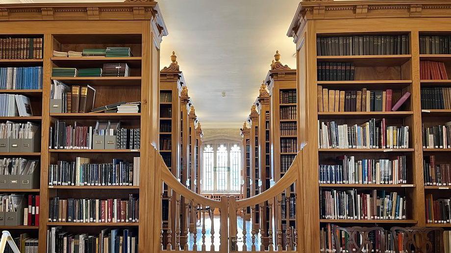 A look down the central walkway of the library's shelves, with a traditional church-style window at the back and a gateway in front. The shelves are very tall and wooden.