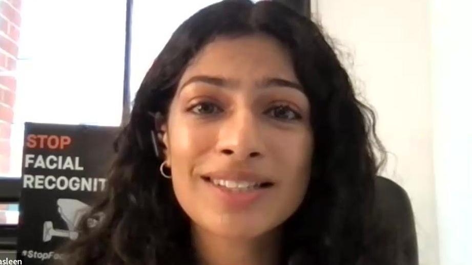 A woman with long dark hair and brown eyes speaks on a Zoom call. There is a poster behind her that says 'stop facial recognition'.