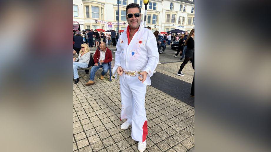 An Elvis impersonator in a white outfit with spots on points at the camera and smiles