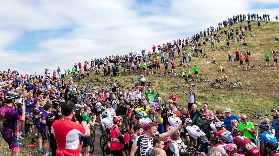 Crowds of cyclists in colourful cycling jerseys and onlookers cheer bike race competitors climbing a steep hill. Onlookers line grassy areas to the side.