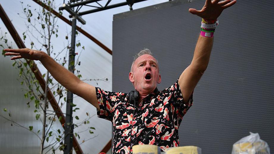 DJ Fatboy Slim plays on stage at Glastonbury Festival. He is stood in front of a darkened screen and has his hands in the air with his mouth open. He has short grey hair and is wearing a black shirt with orange and white koi fish on it. He has a pair of headphones around his neck.