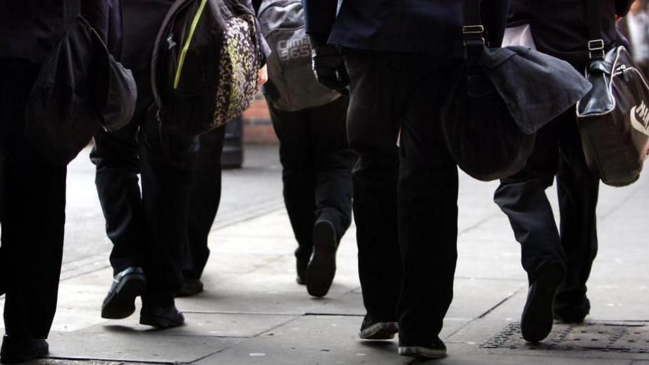 Schoolchildren walking to school, only their feet and legs are in shot. They are wearing black trousers, and black shoes