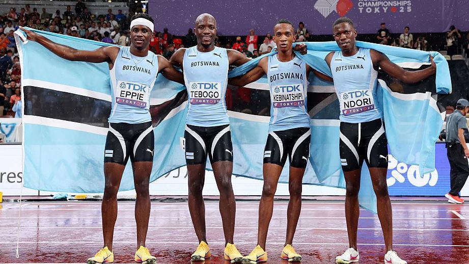 Gold medallists (from left to right) Lee Bhekempilo Eppie, Letsile Tebogo, Collen Kebinatshipi and Bayapo Ndori of Team Botswana pose for a photo on a rainy running track, holding up two Botswana flags behind themselves after winning the men's 4x400m of the World Athletics Championships in Tokyo. All four are wearing black running shorts and sky blue sleeveless Botswana running vests