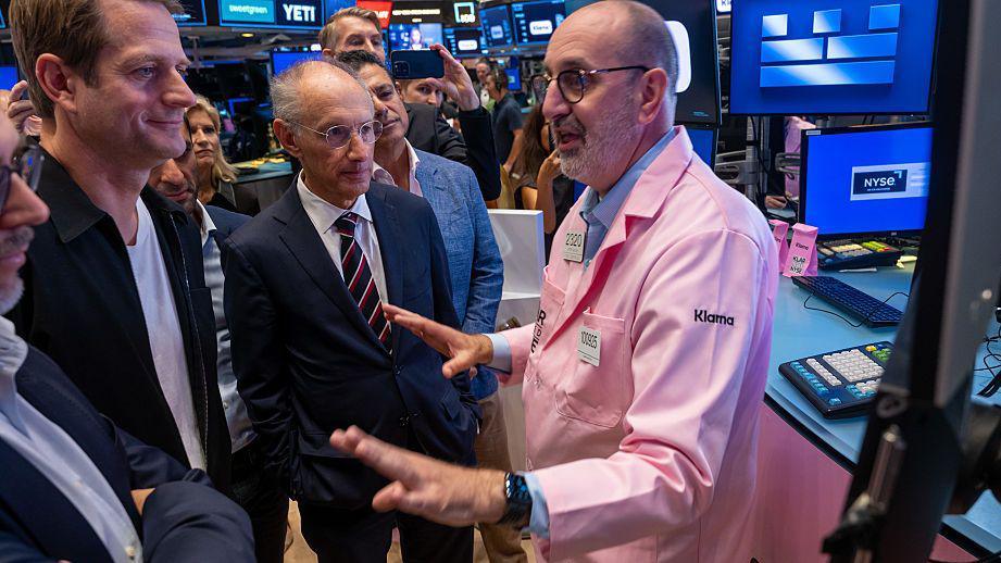 Golygfa ar lawr masnachu’r Farchnad Stoc yn Efrog Newydd, gyda masnachwyr yn sefyll mewn grŵp ger sgriniau digidol. Mae un person yn gwisgo cot binc gyda logo Klarna ac yn esbonio rhywbeth i’r criw o’i flaen, gan ddefnyddio ei ddwy law i bwysleisio. O’u hamgylch, mae sgriniau'n dangos graffiau a logos.