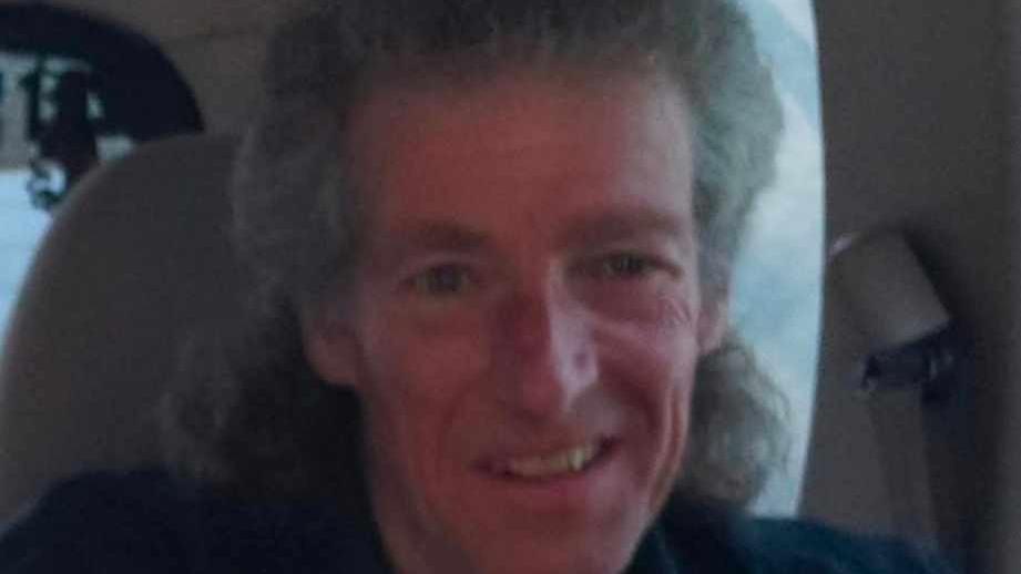 Alban Watts, who has curly grey hair with some of it swept back, smiles at the camera. He is sat in a white seat and is wearing a navy shirt.