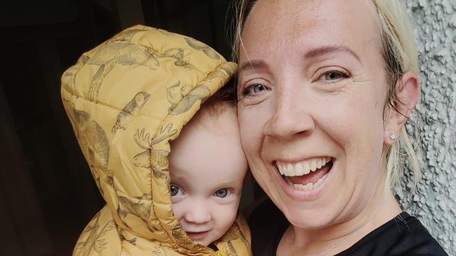 Laura Oakes, who has blonde hair and wears a running top, smiles at the camera while holding her 11-month-old son