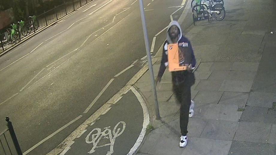 CCTV grab of Marvin Dickson wearing a hooded top and tracksuit trousers and carrying a bright orange pizza box. He is walking at night along a pavement.