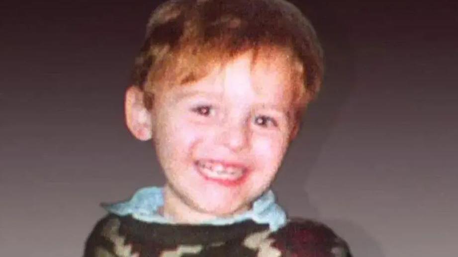 Toddler James Bulger is wearing a brown jumper over a blue shirt and is smiling at the camera.