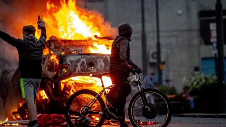 Two youths on a bike with a blazing car in the background