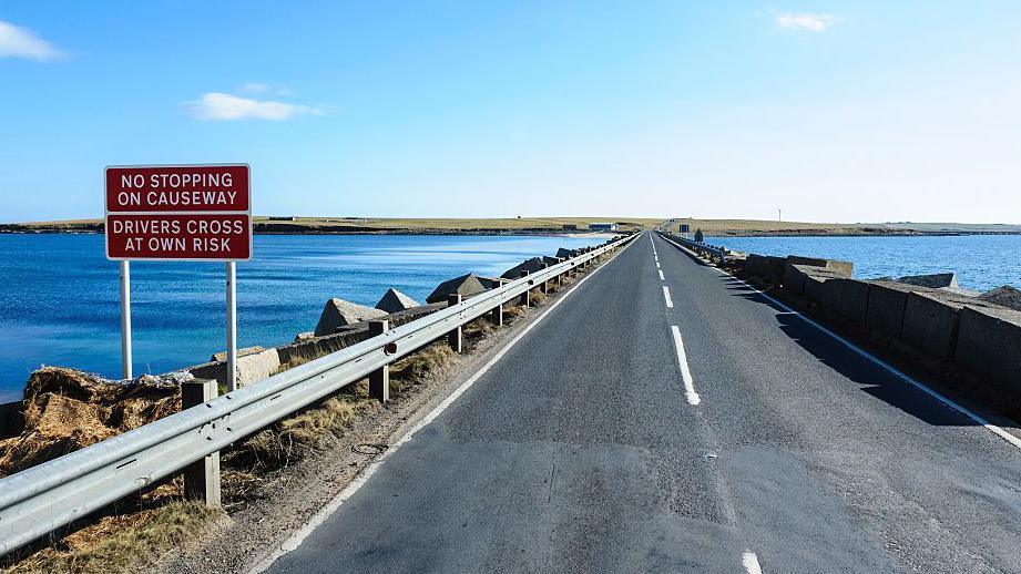 The Churchill Barries at Lamb Holm. The sky and sea are brilliant blue and there is a good quality two track road on the causeway. There is a sign on the left saying "No stopping on causeway. Drivers cross at own risk".