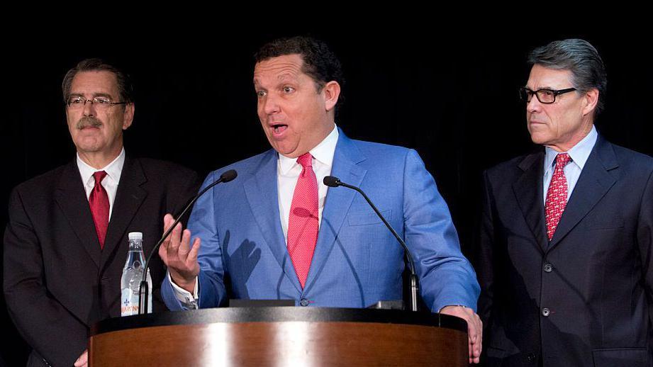 The lawyer Tony Buzbee, wearing a light blue suit, stands at a podium during a press conference, as former Texas governor Rick Perry watches from his left hand side