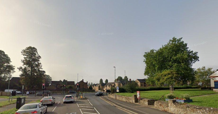 Road junction with traffic lights and cars heading in both directions. A park and a large tree are visible to the right