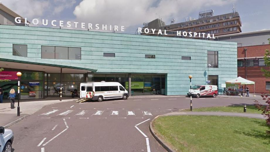 The front exterior of Gloucestershire Royal Hospital. It is a large light blue panelled building with a sign on top. There is an ambulance parked outside with its rear doors open. There is a grassy roundabout on the right and a pedestrian crossing straight ahead.