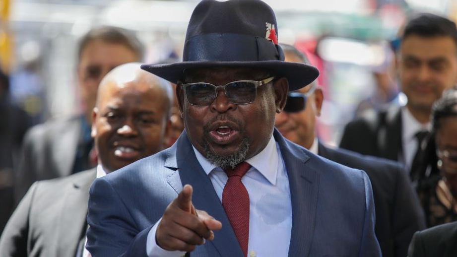 South Africa's finance minister Enoch Godongwana is seen pointing a finger as he prepares to table the national budget. He's wearing a dark blue suit, lighter blue shirt and red tie and has his hat and glasses on.