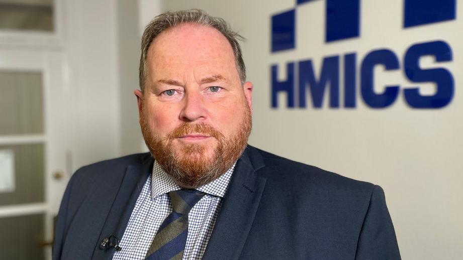 A man with short greying hair and a ginger beard looks at the camera. He is wearing a dark blue suit, chequered shirt and blue and green tie. He is standing in front of a blue HMICS sign.