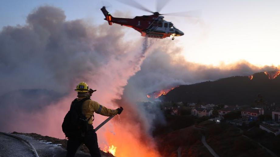 A firefighter douses water on the flames on a hillside above Los Angeles as a helicopter passes over