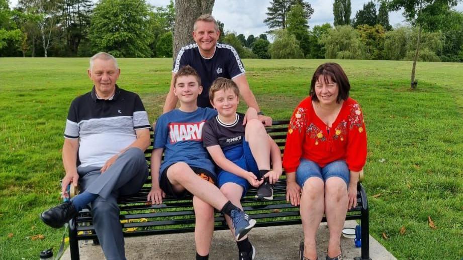 A family photo. Pictured left to right are Kyle's grandad, his older brother, Kyle and his mum. Standing behind the bench is his dad Mark. They are in a park with a field and trees seen behind them.