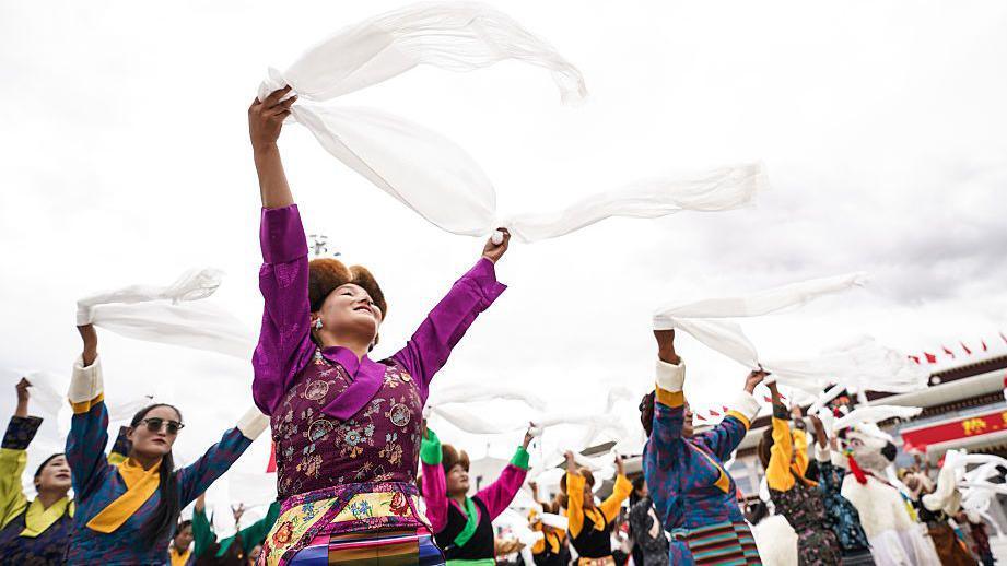 A row of dancers on traditional Tibetan garb hold white cloth into the air