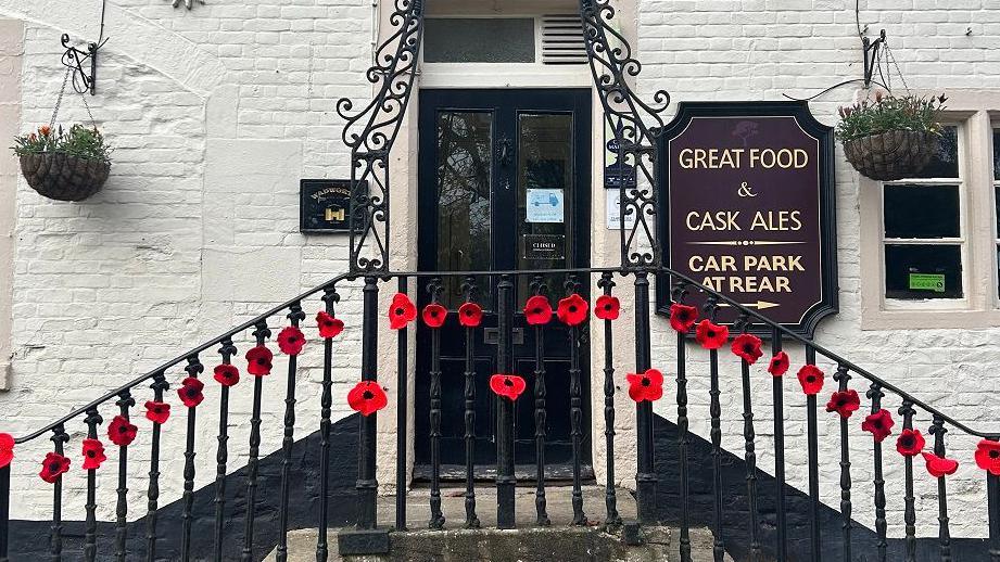 Front of a pub - black door in the middle with steps up to either side, poppies going up and down the railings.