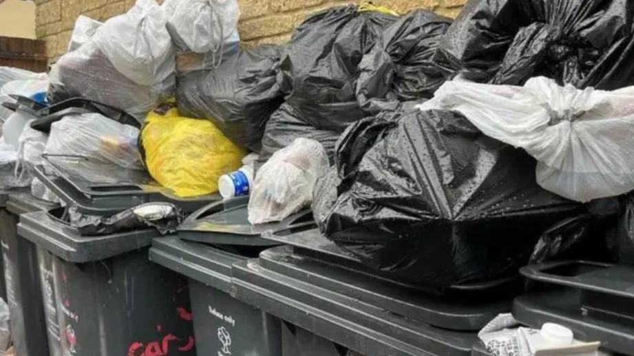 A close-up of several black bins, bursting with rubbish with full bags also laid on top