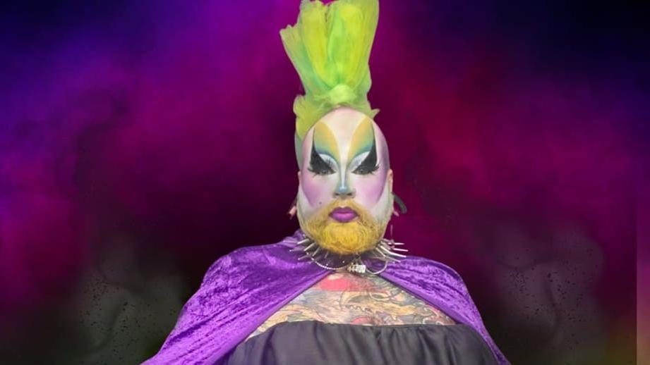 Phil Harper in his drag attire. He has green hair, a purple cape, and purple and green face make-up on. 