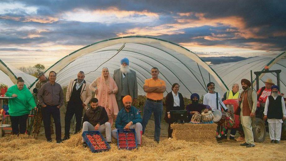 An image from the film showing workers standing in front of polytunnels used to grow strawberries. Some of the Punjabi workers are wearing Indian clothing.