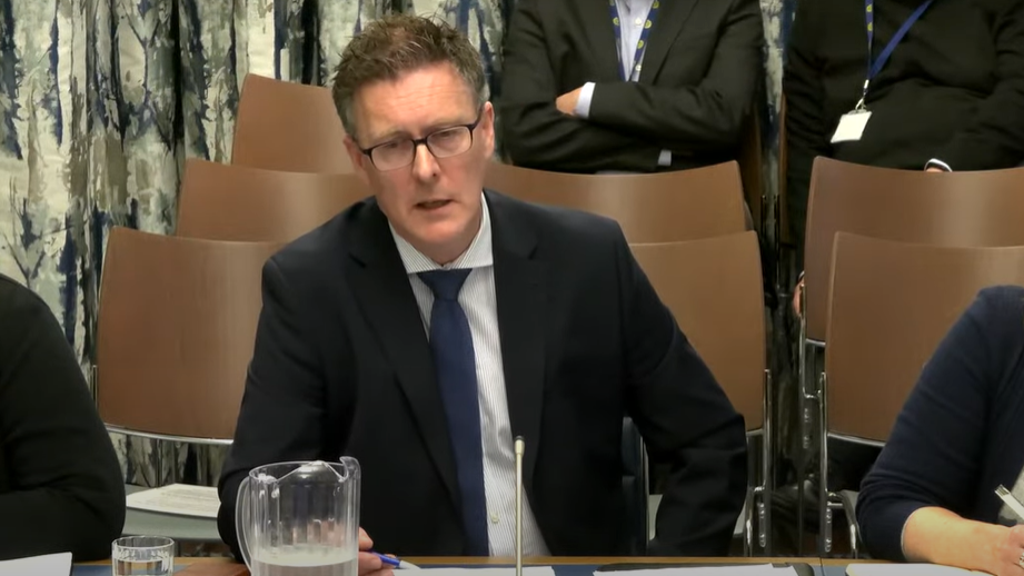 Dr Andrew Brown, sitting in the education committee. He has spikey brown hair and wide glasses and wears a suit. 