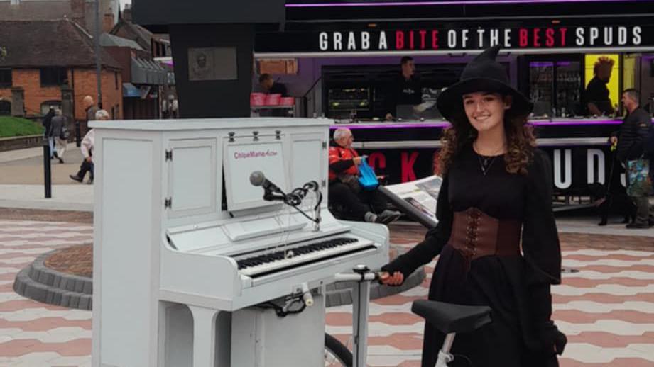 Ms Aston is wearing a witch outfit and is standing next to a white piano on a bike. A microphone is attached to the instrument. She is standing outside Spudman in Tamworth.