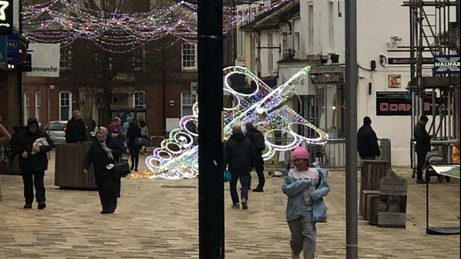 A pedestrian street in Littlehampton with a fully-lit large Christmas tree fallen to the ground. People are continuing to walk in both directions along the pavement