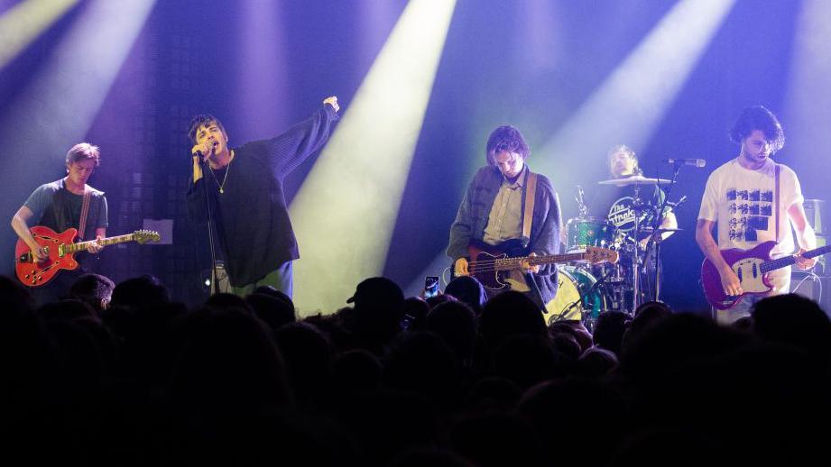 Five men are on a stage. Three play guitar, one sings and one is seated behind a drum kit. Behind them is a purple screen and white strobe lights. 