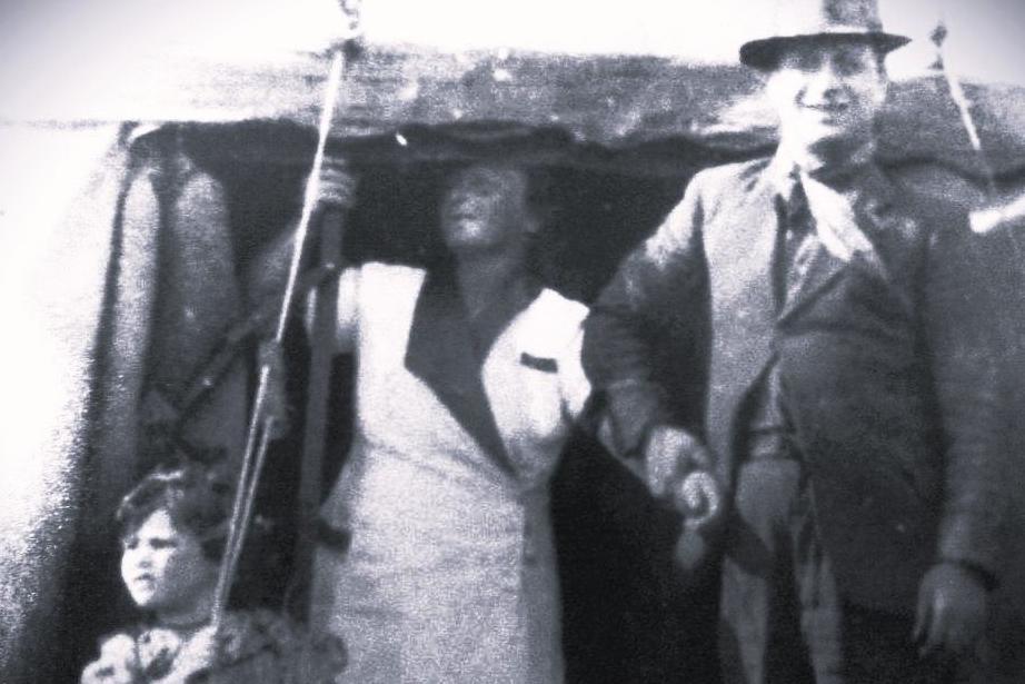 A man wearing a suit standing next to a woman in a white outfit and a child as they set up a tent.