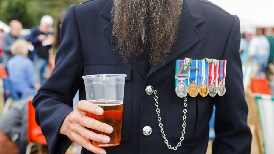 A man holds a pint of beer, he has five gold and silver medals attached to his navy blue blazer on the left side of his chest. He has a long beard. You cannot see the man's face.