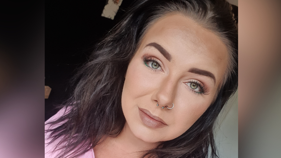 A selfie of Sammi Hill, a 33-year-old woman with dark brown hair. She is wearing a full face of make up and a pink t-shirt. She is subtly smiling.