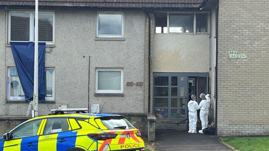 Forensic officers in white suits outside a building, with a police car in the street.