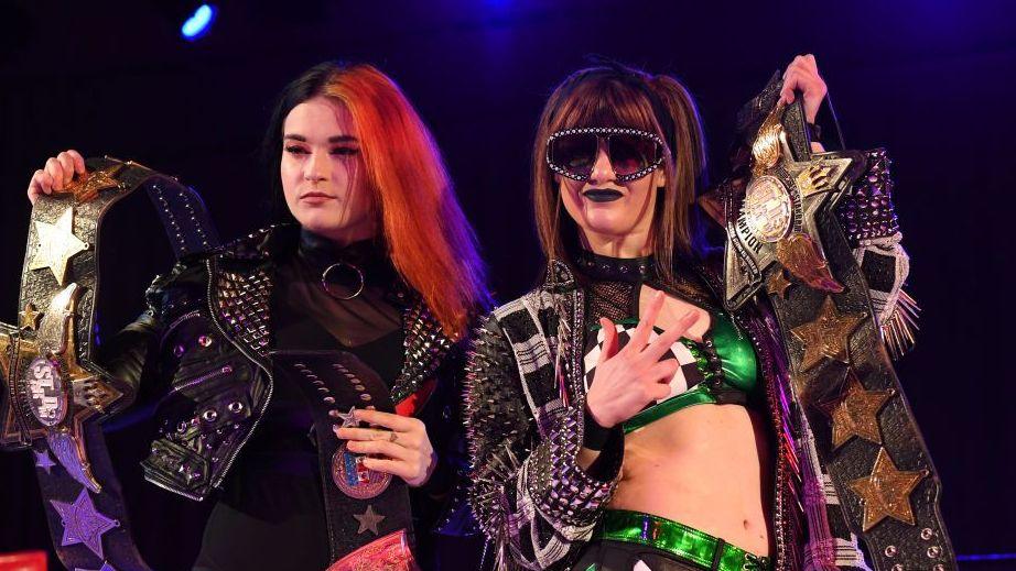 Wrestling tag team Bea Priestley and Jamie Hayter, holding various title belts up ahead of a match. Priestley has long black and orange hair, and is wearing a leather jacket. Hayter is wearing sunglasses, a green and black ring outfit and is also holding up a title belt.
