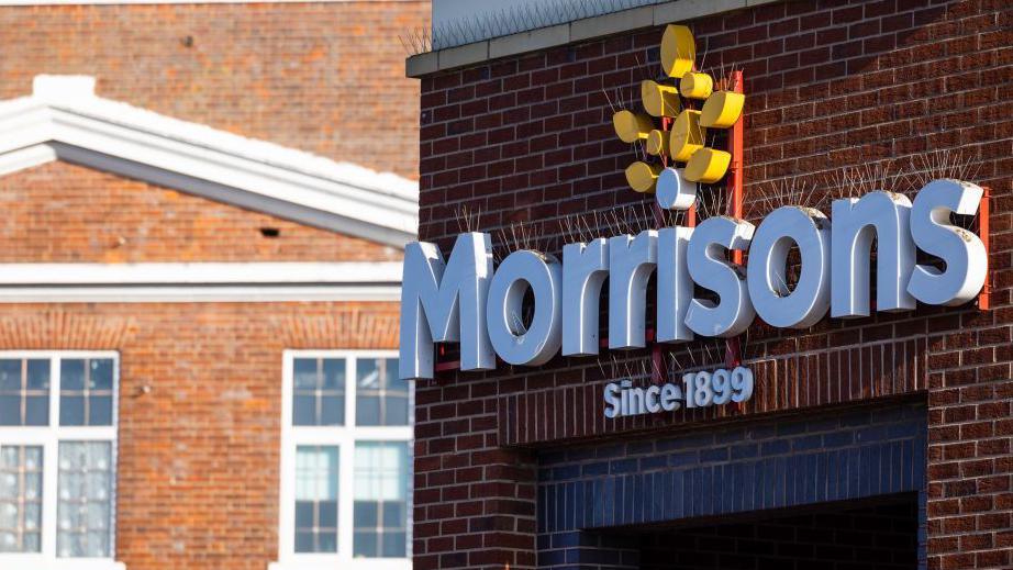 The exterior sign of a Morrisons supermarket, white text with a yellow corn logo, on brick wall