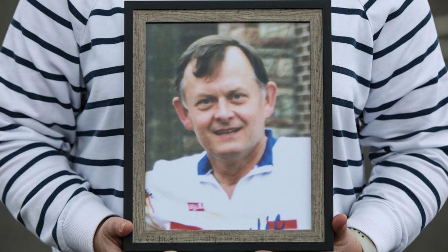 A photo of Sean Brown being held up in a woman's hands. Sean Brown has dark hair and is wearing a white polo shirt. It is in a brown frame and the woman holding it up is wearing a white jumper with thin navy stripes. Only her torso and hands are visible.