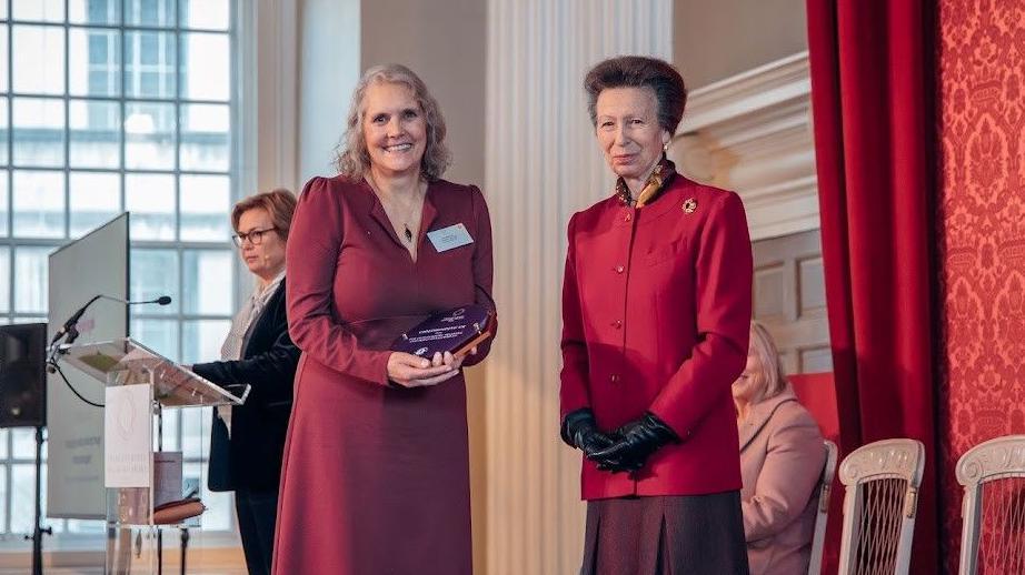 A woman looking at the camera in a red dress holding an award. She is smiling and stoop next to Princess Anne who is wearing a red jacket and black gloves.