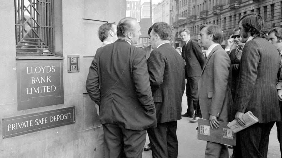 A group of men wearing suits with shaggy hair congregate around Lloyds Bank