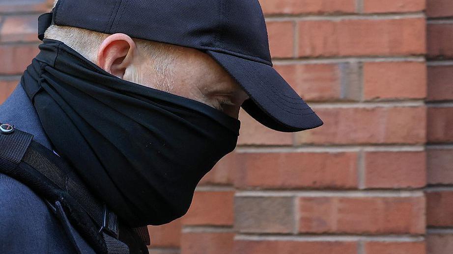 A close-up of the head and shoulders of a man with his face obscured by a black scarf and black cap as he walks towards a red-brick court. His eyes can be seen and his is looking downwards. He is wearing a blue suit jacket and the strap of a bag over his shoulder.